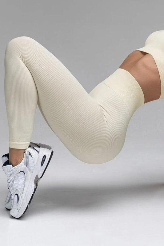 Mulher vestindo uma calça legging canelada bege de cintura alta, em pose fitness, realçando a modelagem justa e o tecido sem costura. O look é combinado com tênis esportivos brancos, transmitindo conforto e estilo para treinos e atividades físicas! Loja nuudi Store nome do produto Calça Legging Fitness Feminina Canelada com Cintura Alta Nuudi Store